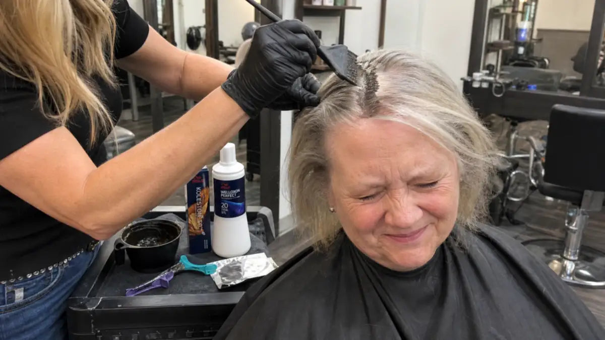 Stylist brushing permanent hair color onto a wincing older woman’s roots at the salon